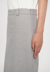 Gros plan d'une personne portant une jupe gris clair avec passants pour ceinture et une fermeture éclair latérale, la main reposant à côté de la jupe.