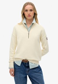 Crème kleurige trui met een ritskraag, geribbelde manchetten, gedragen over een lichtblauw gestreept overhemd met kraag, gecombineerd met donkerblauwe jeans.