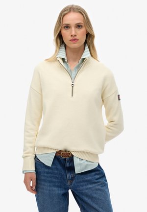 Pull crème avec un col zippé, des poignets côtelés, porté sur une chemise à col rayée bleu clair, associé à un jean bleu foncé.