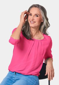 Rosa Bluse mit Dreiviertelärmeln, verziert mit Falten am Halsausschnitt, kombiniert mit hellblauen Jeans. Schlichte, weiche Stofftextur.