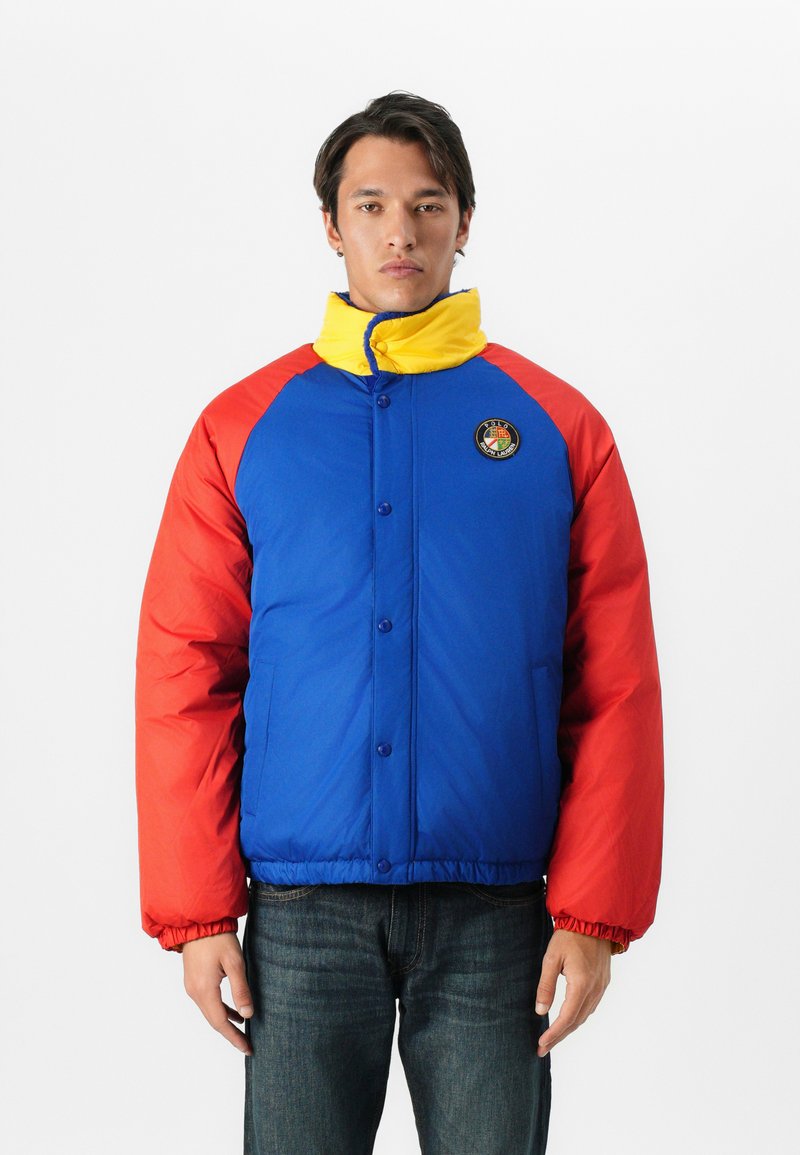 Kolorowa kurtka puchowa z niebieską koronką, czerwonymi rękawami i żółtym kołnierzem. Zawiera logo na klatce piersiowej oraz zapięcie na guziki z elastycznymi mankietami.