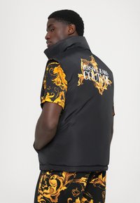 Gilet rembourré noir avec col montant, présentant un motif floral doré au dos et sur l'épaule, marqué "Versace Jeans Couture" en texte blanc.