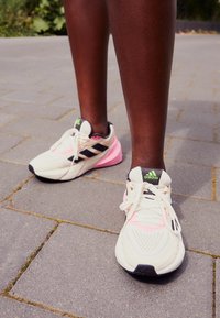 Baskets de course beiges avec des accents noirs et roses, dotées de dessus en mesh, de semelles épaisses et de lacets blancs, posées sur une surface carrelée grise.