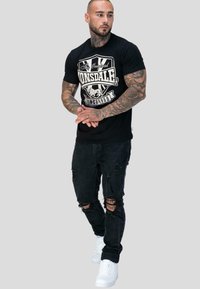 Mies, jolla on tatuointeja, pukeutuneena mustaan Lonsdale-t-paitaan, revenneisiin mustiin farkkuihin ja valkoisiin tennareihin, seisoo tasaisella harmaalla taustalla.