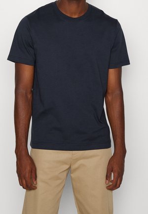 Mann trägt ein schlichtes dunkelblaues T-Shirt mit kurzen Ärmeln und beige Hose, steht mit entspannt an den Seiten hängenden Armen vor einem schlichten Hintergrund.