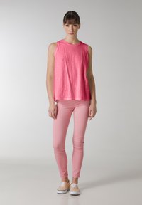 En rosa ärmlös topp med ljusrosa tajta byxor och beige skor. Toppen har en strukturerad yta; byxorna är släta och smala.