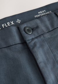 Ceinture de pantalon gris foncé avec fermeture par bouton et étiquette intérieure visible indiquant "FLEX" et "NEXT PERFORMANCE."