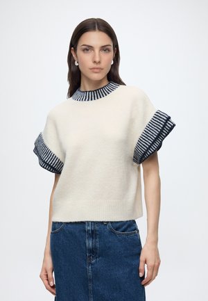 Donna con capelli scuri che indossa un maglione bianco a maniche corte con polsini e colletto a righe blu navy, abbinato a una gonna di jeans blu.
