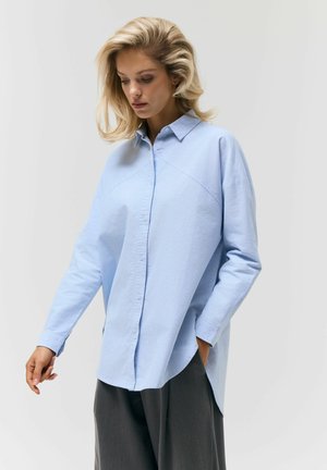 Kobieta o blond włosach, ubrana w luźną jasnoniebieską koszulę na guziki i ciemne szerokie spodnie, patrząca w dół, z jedną ręką w kieszeni.