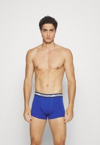 Blue cotton boxer shorts with a black waistband featuring the brand name repeated. The model is shirtless, standing upright with arms relaxed.