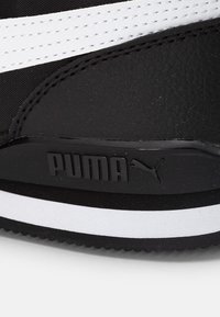 Scarpa sportiva nera con superficie testurizzata, strisce bianche e un logo Puma nero ben in vista sul lato. Suola in gomma visibile.