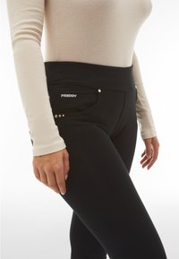 Leggings neri con un ampio cinturino in vita, caratterizzati da accenti metallici e un logo. Il tessuto sembra elastico e liscio. Si indossa un top aderente a coste.