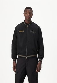 Black bomber jacket with gold accents, zip closure, ribbed cuffs, featuring embroidered text and logo on the left chest area.
