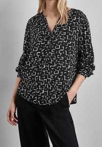 Svart blus med ett vitt geometriskt mönster, V-hals, lång ärm med rynkade manschetter och böjd fåll, tillverkad av lätt tyg.
