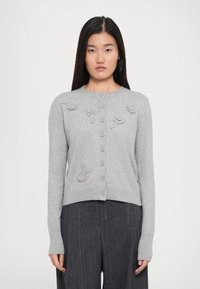 Donna che indossa un cardigan grigio chiaro con bottoni e dettagli in maglia floreale, abbinato a pantaloni grigio scuro a righe, in piedi su uno sfondo bianco semplice.