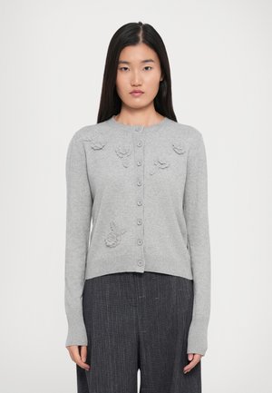 Kvinne iført lys grå knappet cardigan med blomsterstrikkdetaljer og mørkegrå pinstripede bukser, stående mot en enkel hvit bakgrunn.