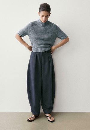 Kobieta poprawiająca sweter z krótkim szarym rękawem, ubrana w luźne ciemne spodnie i czarne sandały, stojąca na tle gładkiej białej ściany.