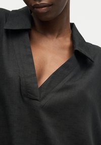 Gros plan d'une personne portant une chemise noire à col avec un profond décolleté en V, montrant le cou et la partie supérieure de la poitrine.