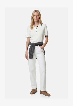 Kobieta ubrana w biały polo z krótkim rękawem i czarnym lamówką, białe spodnie, brązowy pasek, brązowe mokasyny oraz szary sweter związany w talii.