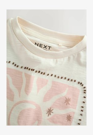 T-shirt blanc cassé avec un motif de soleil rose et d'étoile, décoré de petits clous métalliques le long du bord supérieur et une étiquette de col affichant "NEXT".