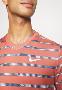 Camiseta deportiva con cuello en V en color coral con rayas horizontales en azul marino y azul claro. Presenta un logo blanco de Nike en el pecho. Tela suave y ligera.