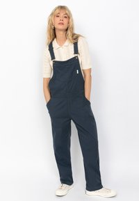 Marineblauwe overalls van duurzaam materiaal, met een ontspannen pasvorm, voorzakken, verstelbare banden en gouden hardware-accenten.