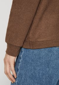 Felpa marrone con polsini e orlo a costine, abbinata a jeans in denim blu chiaro. La vista ravvicinata mette in evidenza la texture e i dettagli del tessuto.
