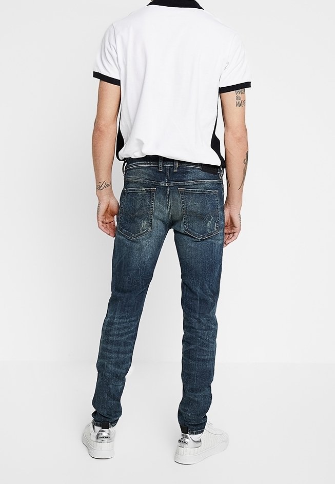 Man i vit och svart polotröja, mörkblå slim-fit jeans och vita sneakers, stående vänd bort mot vit bakgrund.