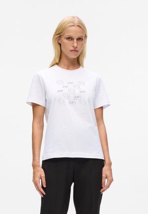 Mujer rubia vestida con una camiseta blanca con pequeñas caras contorneadas y pantalones negros, de pie frente a un fondo blanco liso.