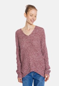 Bordeauxfarbener Strickpullover mit V-Ausschnitt und überschnittenen Schultern, ausgestattet mit einem strukturierten Muster und seitlichen Schlitzen am Saum. Getragen mit blauen Jeans.