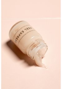 Glazen pot met lichtbeige crème met een licht ondoorzichtige textuur, gelabeld met zwarte tekst. Crème morst uit de pot op een roze oppervlak.