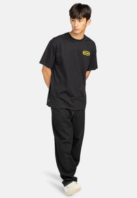 Black cotton t-shirt with short sleeves, featuring a yellow rectangular logo on the left chest. Paired with loose black jeans and white sneakers.