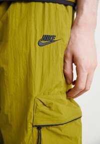 Nike olive green pants with a textured finish, featuring a black logo and a side pocket with drawstring closure.
