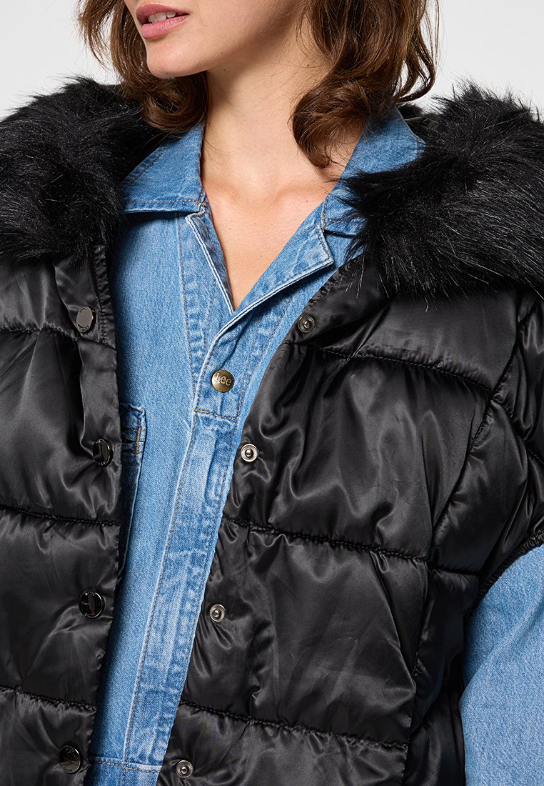 Gilet imbottito nero con colletto in pelliccia sintetica e chiusura con bottoni a pressione, abbinato a una camicia di jeans azzurro chiaro, che mette in mostra una texture liscia e un design a strati.