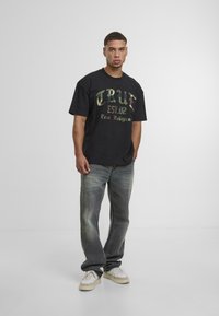 Camiseta de algodón negra con texto multicolor, "TRUE EST.02 True Religion." Combinada con jeans grises desgastados y zapatillas de color claro.