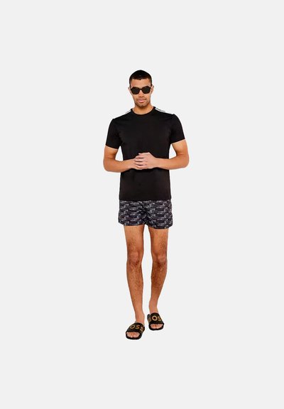 T-shirt noir à manches courtes, shorts de bain à motifs dans des tons sombres, sandales noires avec logo doré, se tenant devant un fond uni.