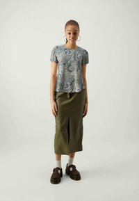 Frau trägt ein Kurzarmoberteil mit Paisley-Muster, einen olivgrünen Midirock mit Schlitz vorne, leichte Socken und dunkelbraune Plateauschuhe.