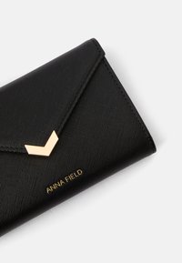Carteira sintética preta com uma superfície texturizada, apresentando um detalhe em chevron dourado e texto em relevo "ANNA FIELD". Design slim e retangular.