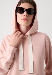 Persona che indossa un hoodie rosa pallido con ampi lacci bianchi e occhiali da sole a forma di gatto neri, tenendo il cappuccio vicino al viso.