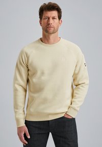 Sweatshirt beige à col rond avec des poignets et un ourlet côtelés, présentant un petit logo sur la poitrine et un accent à boutons sur la manche.