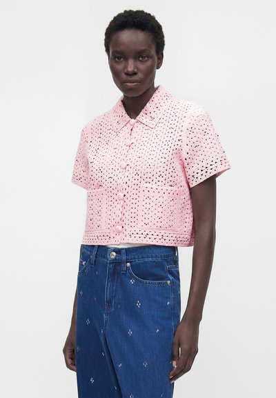 Γυναίκα με σκούρο δέρμα που φοράει ροζ πλεκτό τοπ με κουμπιά και κοντά μανίκια και μπλε τζιν ψηλόμεσα με μικρή λευκή κεντητική.