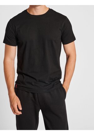 Homme portant un t-shirt noir uni à manches courtes et un pantalon noir, une main dans la poche, debout devant un fond clair.