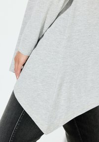Helle graue, strukturierte Tunika mit langen Ärmeln und abgerundetem Saum, verziert mit gelben Nähten, kombiniert mit schwarzen eng anliegenden Jeans.