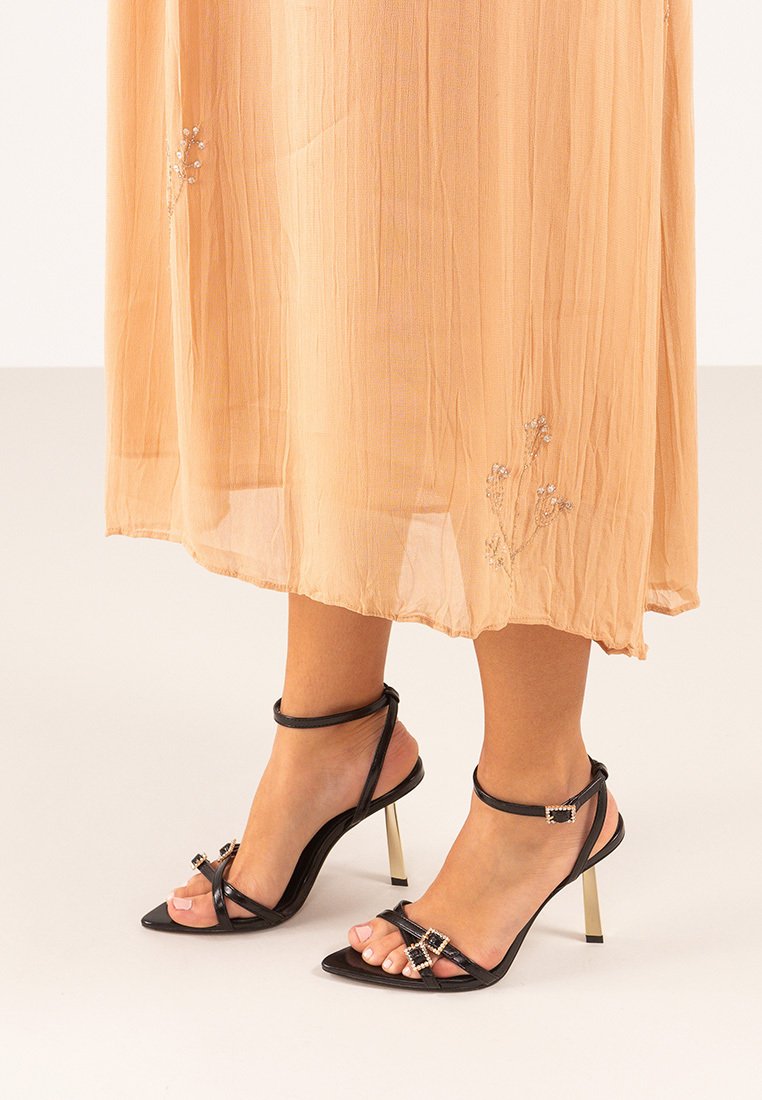 Los tacones de aguja negros tienen un diseño con tiras, correas ajustables en el tobillo y acentos en tono dorado. La suela es elegante y con punta redondeada.
