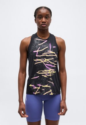 Atlétikus személy áll előre nézve, fekete ujjatlan pólót visel, amelyen rózsaszín és sárga absztrakt vonalak vannak, valamint lila rövidnadrágot.