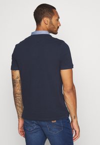 Marinblå pikétröja med krage i chambray, med korta ärmar och en texturerad design. Baksidan visar en diskret logopatch nära fållen.