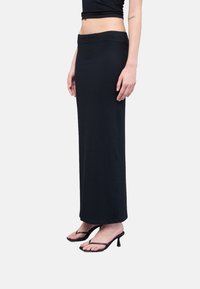 Falda maxi negra de canalé con cintura ajustada, textura suave y abertura lateral, combinada con sandalias de tacón negras con tiras finas.