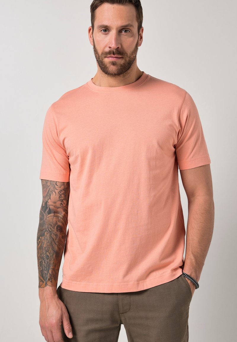 Homme barbu avec un bras gauche tatoué portant un t-shirt à manches courtes pêche et un pantalon marron, debout avec la main droite dans la poche devant un fond clair.