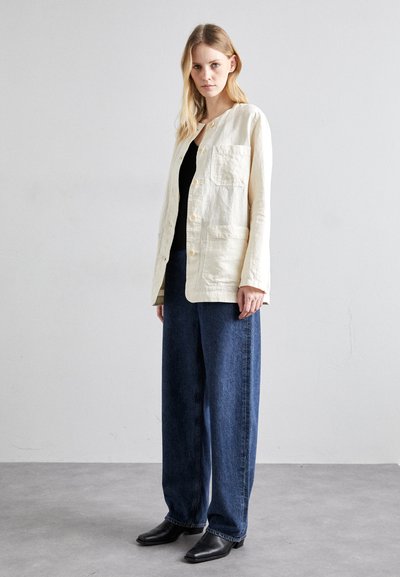 Veste en lin beige clair avec un col rond et des poches avant, associée à un pantalon en denim bleu ample et des bottines noires.