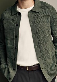 Cardigan testurizzato verde oliva con chiusura a bottoni, colletto a coste e intricati motivi a maglia, indossato sopra una camicia bianca aderente e una cintura marrone.